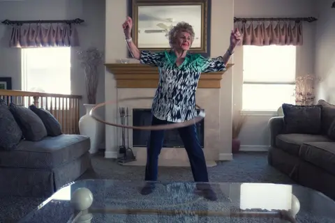 Chris Riess and Amy Hill Joan in a pair of dark denim jeans in an aztec shirt while spinning with a hoola hoop around her waist in a lounge with brown sofa, a grey carpet and a mirror above a mantlepiece