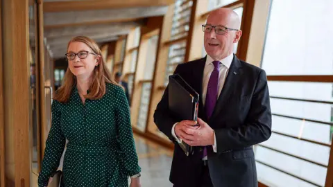Getty Images Shirley-Anne Somerville - a woman with red shoulder length hair in a green dress - and first minister John Swinney - a bald man in a dark suit with purple tie - walking together into the Scottish Parliament chamber