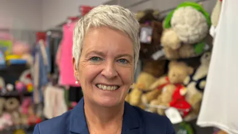 Report author Edel Wilson - a smiling woman with short blonde hair, wearing a blue blazer and drop pearl earrings - stands in front of shelves of soft toys.