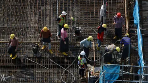 Getty Images Migrant workers, wearing yellow helmets and long-sleeved shirts, stand at a building construction site on 3 May, 2023 in Singapore