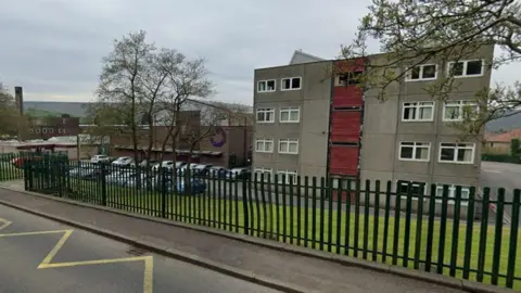 Google External view of The Valley Leadership Academy in Bacup which has a black fence and a handful of trees at the front of the school and car park to the right with hills in the distance. 