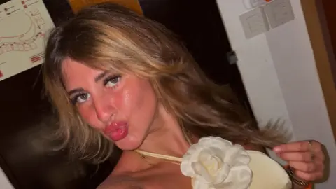 Bella Culley A selfie taken by Bella Culley. She is wearing a beige dress with a white fabric flower at her neck and has on pink lipstick and black mascara. She is pouting at the camera and holding a strand of hair in her hand.