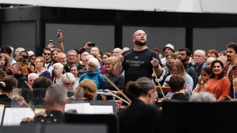 PA Media A conductor wearing a black T-shirt that reads "Birmingham forever" leads a large orchestra playing a concert in a station, while a crowd watches behind him.