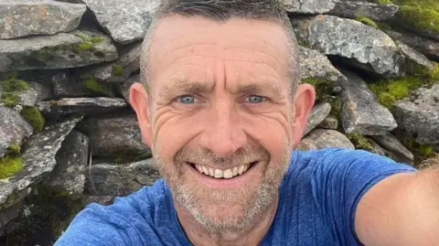 Scott Mitchell family Mr Mitchell has neatly cut grey hair. He has stubble around his mouth and chin and is smiling. He is wearing a light blue running top and is taking a selfie while next to a stone wall.