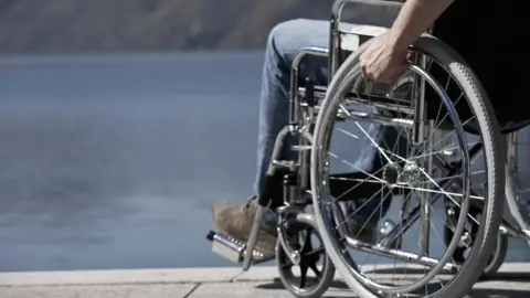 BBC A stock close-up image of a person in a wheelchair showing a man's hand on a wheel rim and a foot in brown suede shoe on a footrest. The wheelchair is on a pavement overlooking a body of water.