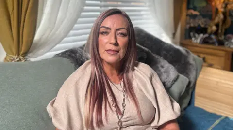 BBC Julieanne Boyle sitting on a green sofa in a living room looking directly at the camera. She has long, straight hair with blonde and copper tones. She is wearing a loose beige top, and a long silver and pearl necklace.