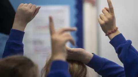 PA Media School children in a classroom. They have their hands up and only their hands, arm and top of their heads are visible. They are wearing blue jumpers and one of the children has a bracelet on.