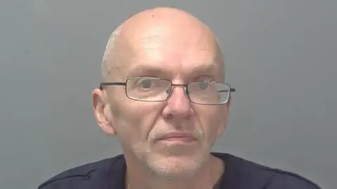 Suffolk Police A mugshot of Roy Barclay - a bald man wearing glasses. He is standing against a grey backdrop and is looking directly into the camera.