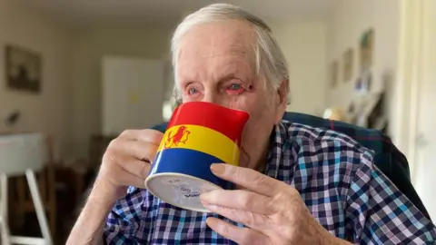 Alex Pope/BBC Leslie Lemon, with a blue, yellow and red custard jug featuring three birds, held to his lips. He is wearing a blue checked shirt and has short grey hair. 