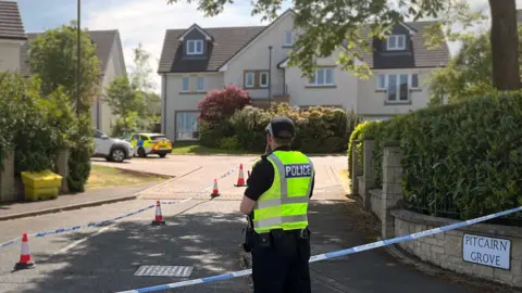 BBC A police officer standing in a hi vis vest in front of a street with a police car