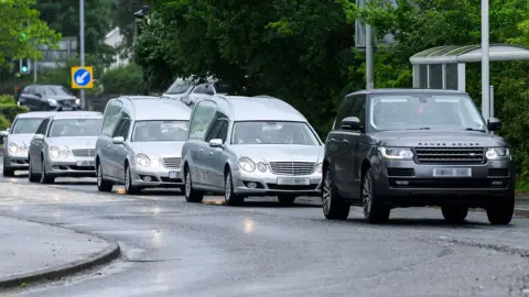 BBC A convoy of silver hearses and mercedes, led by a black Range Rover driving along a road