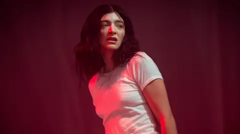 Getty Images Lorde on stage at Glastonbury. The singer, lit in a red light, wears a white T-shirt, her arms by her side. Her dark hair is worn loose and messy. She looks over her left shoulder towards the crowd.