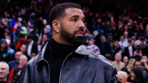 Getty Images Drake pictured at a basketball game. The rapper, who has short hair and a cropped beard, wears a diamond stud earring and a black leather jacket over a black turtle neck jumper. He looks away from the camera over his right shoulder and crowds of spectators are in the background. 