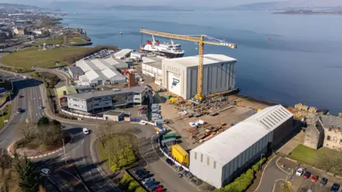 BBC An overhead drone shot of the Ferguson Marine Port Glasgow shipyard. It includes several warehouses and a yellow crane, with cars parked around the edges. It sits on the Clyde river, which can be seen in the background.