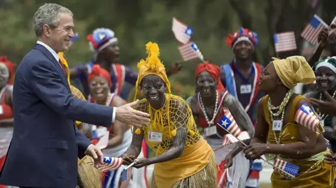 AFP via Getty Images Former US President George W. Bush wearing a black suit reaching out to dancers who are wearing traditional attire and holding Liberian flags