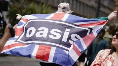 PA Media A man is holding a Union Jack flag which has the black and white Oasis logo in the middle. He is wearing a bucket hat and shorts. 