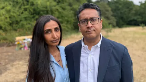 BBC Smera Chohan and Sajjad Butt look towards the camera while standing in front of a wooden bench commemorating their daughter Nuria.