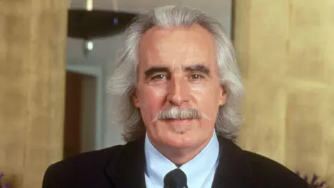 Charlie Miller Salons Charlie Miller has shoulder-length silvery hair and moustache. He is wearing a black suit jacket, white shirt and tie.
