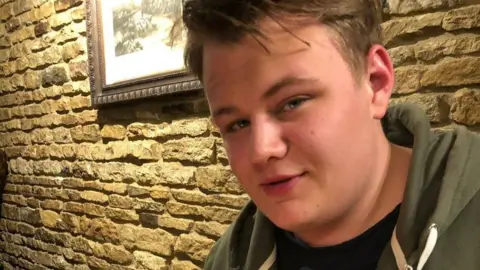 PA Media Harry Dunn had fair hair and was wearing a khaki green hoodie and black T-shirt. Behind him is a stone wall with a framed picture.