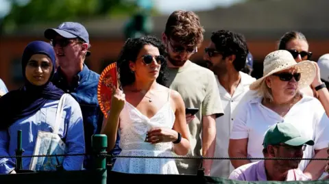PA Media Spectators stand at Wimbledon - with one woman with curly dark hair and a white top standing in the middle holding a fan to cool down from the heat.