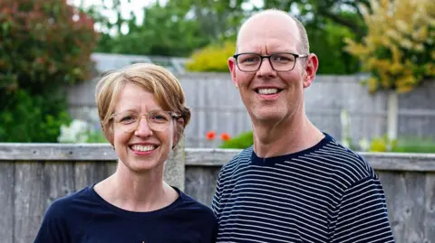 Family Handout/PA Wire A smiling Chris Marriott with his wife Bryony stands in front of a wooden garden fence in a striped T-shirt and glasses