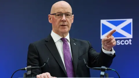 Getty Images A bald man with glasses points with his left hand while speaking at a podium. He is wearing a dark suit, white shirt and purple tie. There is a blue background behind him with a saltire on it 