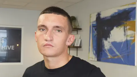 BBC Image of a young man in a black t-shirt, staring directly into the camera. He has short cropped black hair and is clean shaven. He is stood in a room with a large abstract blue, white and black painting behind him, and a shelf with smaller objects on it. 