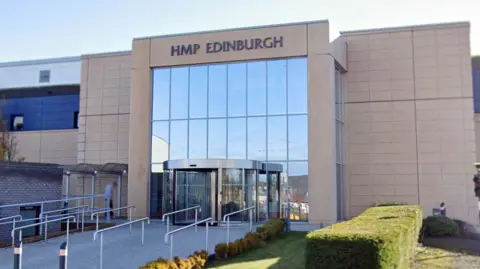 Google The entrance facade to a prison building, a large glass front and modern beige brick panels above a revolving door. A sign at the top says HMP Edinburgh