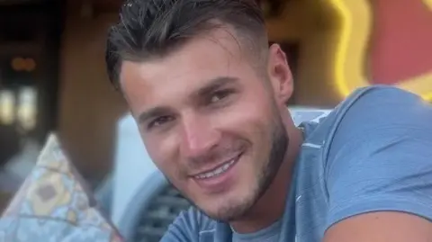 Met Police Blue Stevens is wearing a blue t-shirt smiling at the camera. He has short brown hair and slight stubble.