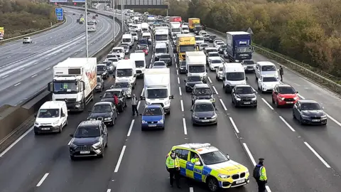 Just Stop Oil/PA Media Six lanes of traffic on motorway have all been stopped with a police car parked in the centre. The cars are facing the camera and the image has been taken from above the M25.