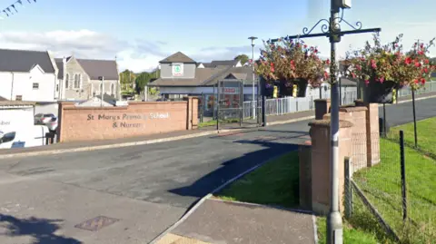 Google An image of the school gates of St Mary's Primary School. There are two red bricked walls leading into the main entrance. On the red wall a sign reads "St Mary's Primary School & Nursery". A Spar shop can be seen in the background.