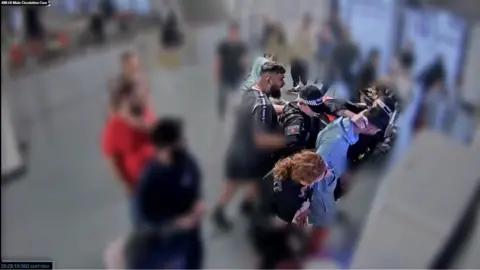PA Media Police officers surround a man in blue at an airport pay station. One has a hand on the back of his neck. A female officer with long red hair holds his arm