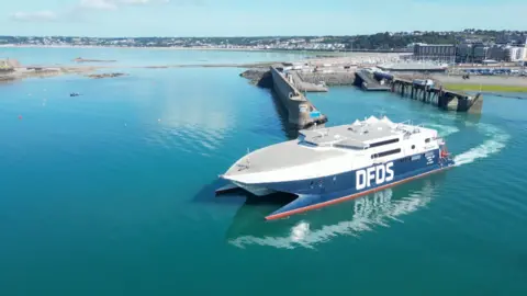 BBC A DFDS ferry departs St Helier in Jersey on a sunny day. The water appears to be still. 