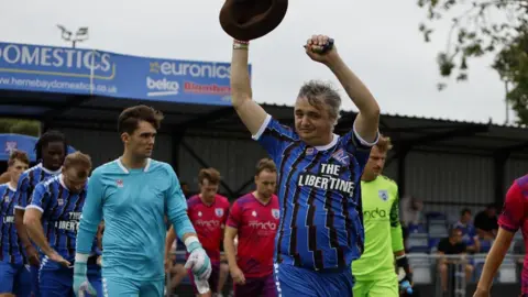 Margate FC Pete Doherty raising his arms and a brown hat to the crowd as he leads out a football team in blue and black kits, which he is also wearing.