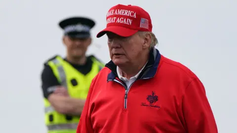 PA Media A man dressed in a red jumper and red hat, with the logo "make America great again" on it. In the background, a police officer stands with his arms folded. He is wearing a reflective vest and large black hat. 