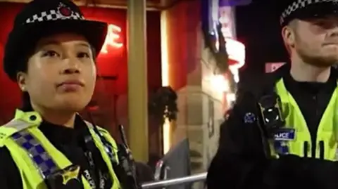 GMP Two uniformed police officers, one male and one female, stand in front of the neon signs of a Manchester nightspot