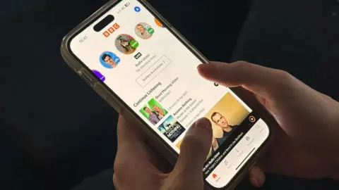 BBC Hands with pink nail polish hold an iPhone with the BBC Sounds app on the screen. 