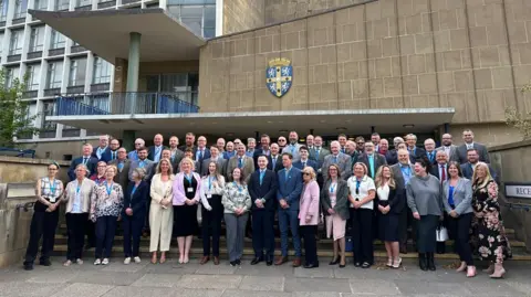 BBC The new council members smiling as they line the steps in front of Durham County Hall in May 2025. It is a beige building with a lot of windows and the county crest.
