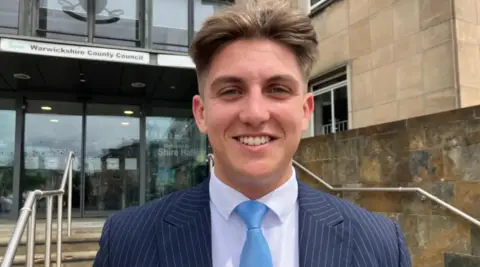 BBC A young man with short light brown hair is wearing a blue pinstriped suit, a white shirt and pale blue tie. In the background are steps leading up to a large glass building with a sign above double doors that reads, "Warwickshire County Council".