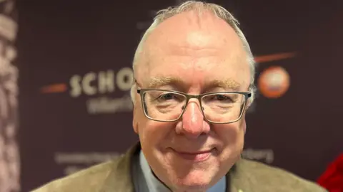 BBC Mervyn Gibson smiling in a beige suit with short white hair and glasses. 