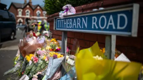 PA Media A row of flowers lay next to a small brick wall and a sign reading 'Tithebarn Road'