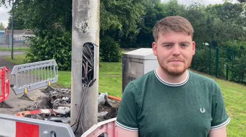 BBC A man in a green shirt is stood in front of a metal lamp post. There is a hole cut out of the post with thick wiring protruding from the hole, it is scorched and badly damaged.