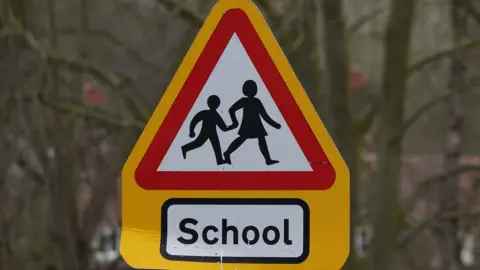 BBC A yellow triangle 'children crossing' sign with 'School' below the sign in black text.
