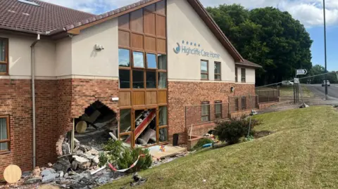 BBC A large hole at the bottom left of a brick and white stone building next to a window. Bricks and debris from the wall are scattered all around and in the bushes and grass in front of the building. 