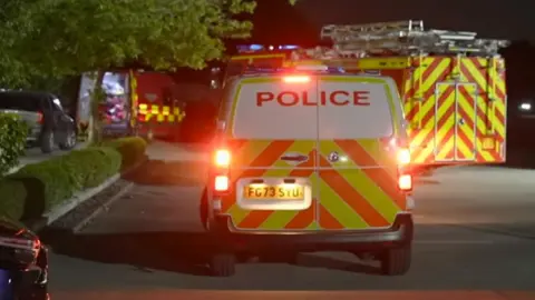 BBC The rear of a police van in a car park with yellow and orange chevrons on the back and its lights flashing with a fire engine behind it