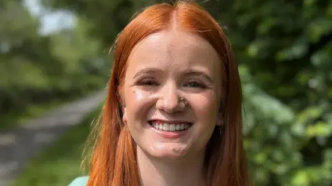 BBC Eve has long ginger hair and is smiling at the camera. She is outdoors, with a pathway and hedges blurred in the background. She is wearing a pale green top.