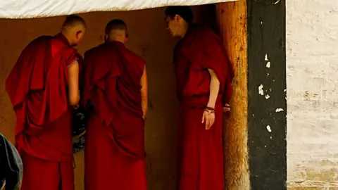 Xiqing Wang/ BBC Three monks stand in a row in red robes talking among themselves, their faces truned away from the camera
