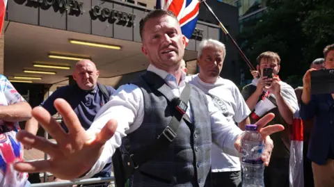 PA Media Tommy Robinson is wearing a light blue shirt and a checked blue waistcoat with a messenger bag over his shoulder. He is speaking with his hands open and outwards while holding a water bottle. A crowd of men surround him outside court.