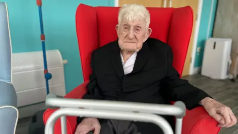 BBC Donald Rose with short grey hair in a black blazer and white shirt sat in red armchair.
