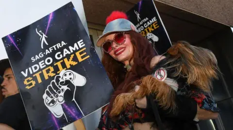 Getty Images A costumed performer wears a fake fur coat and red-tinted sunglasses as she carries a placard with the words "Sag-Aftra video game strike" and a picture of a hand clutching a gamepad printed on it.